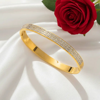 A gold bangle bracelet with two rows of small sparkling stones resting on smooth white satin fabric, with a red rose positioned beside it.