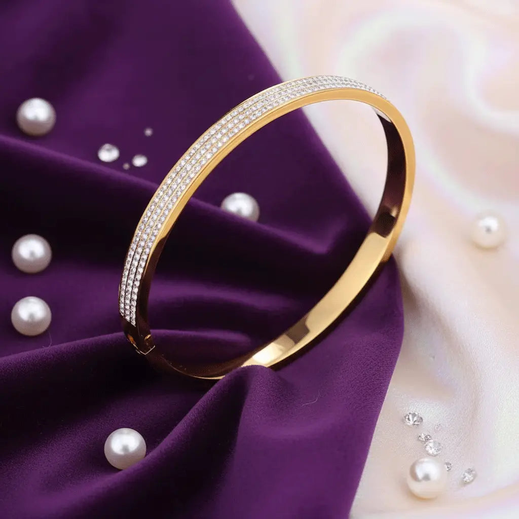 A gold bangle bracelet with two rows of small sparkling stones resting on smooth purple satin fabric, with a pearls positioned beside it.
