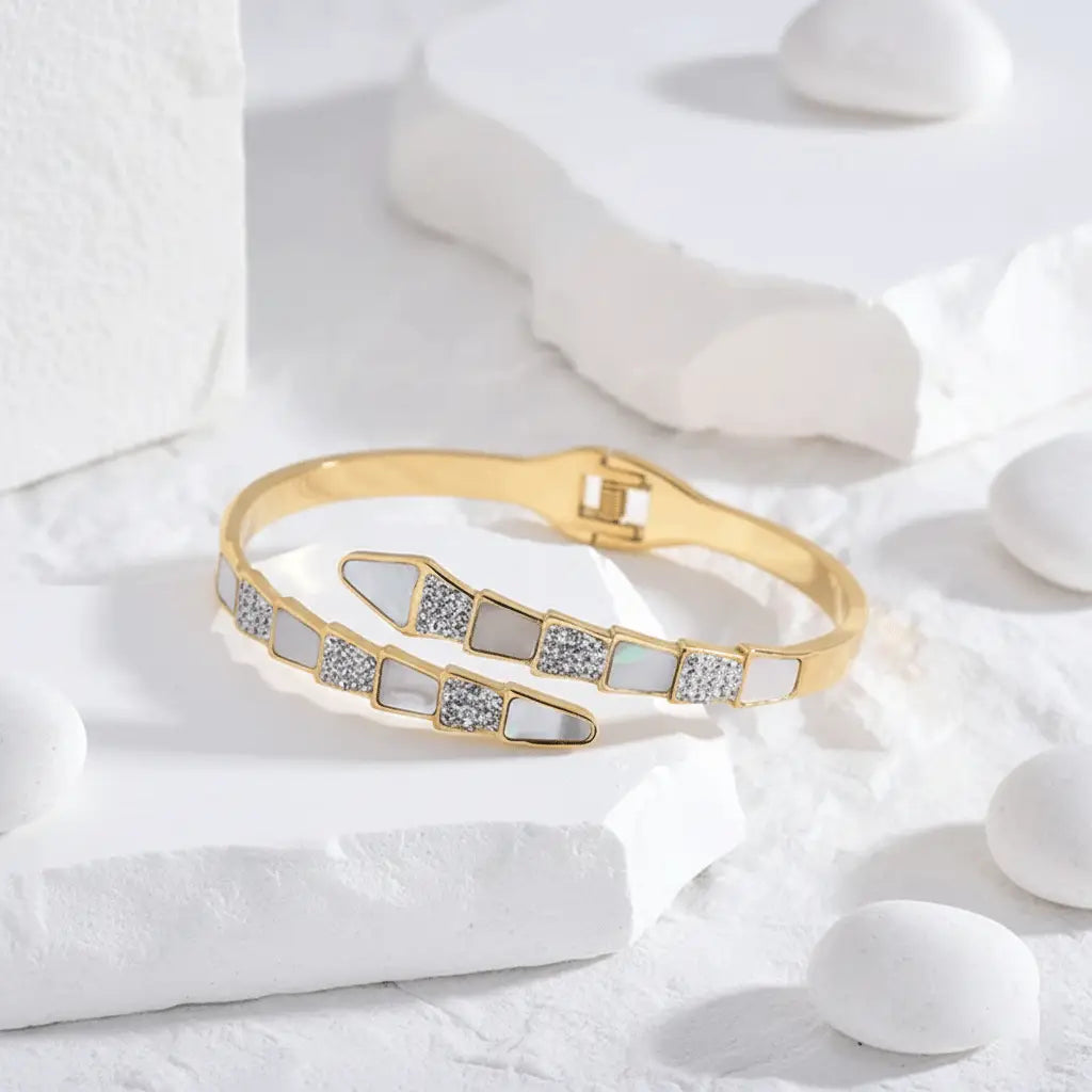A gold snake bracelet featuring a modern geometric design with alternating triangular segments of white enamel and sparkling crystals, displayed on a white textured surface .