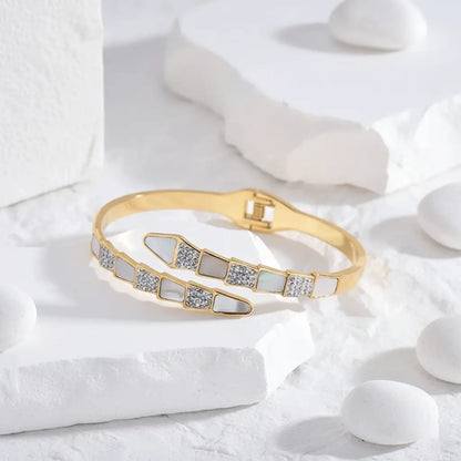 A gold snake bracelet featuring a modern geometric design with alternating triangular segments of white enamel and sparkling crystals, displayed on a white textured surface .