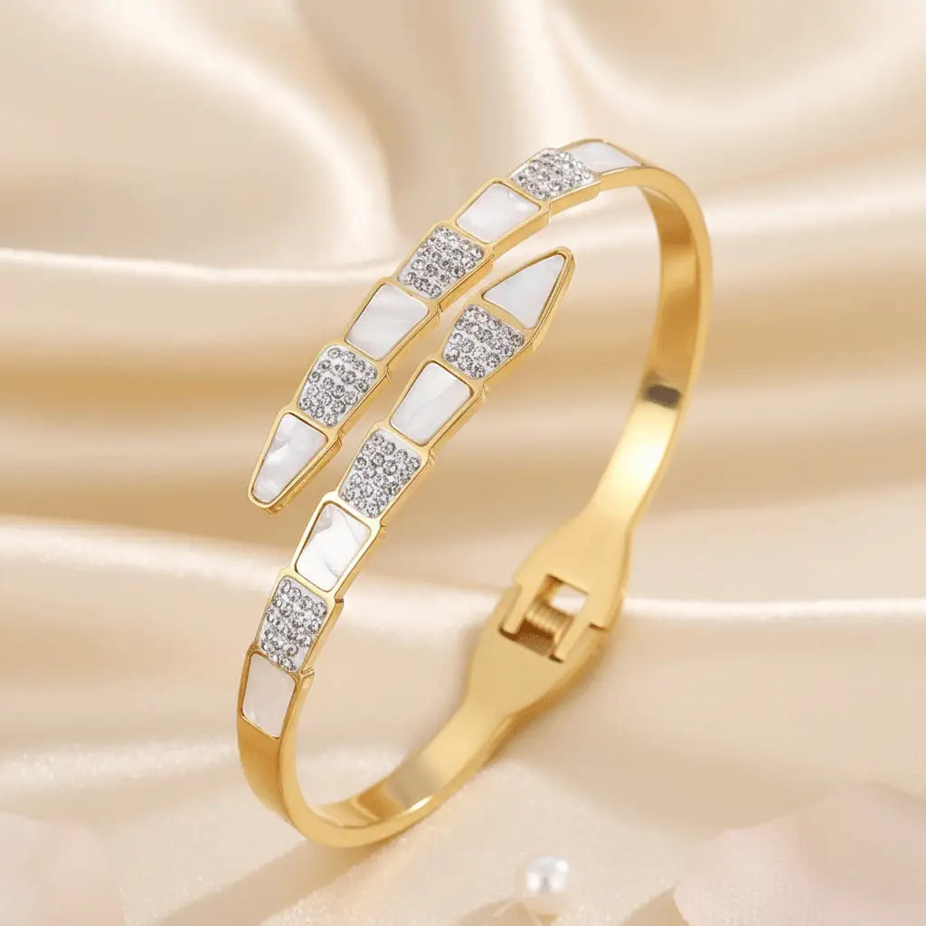 A gold snake bracelet featuring a modern geometric design with alternating triangular segments of white enamel and sparkling crystals, displayed on a satin cloth.