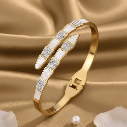A gold snake bracelet featuring a modern geometric design with alternating triangular segments of white enamel and sparkling crystals, displayed on a light brown cloth.