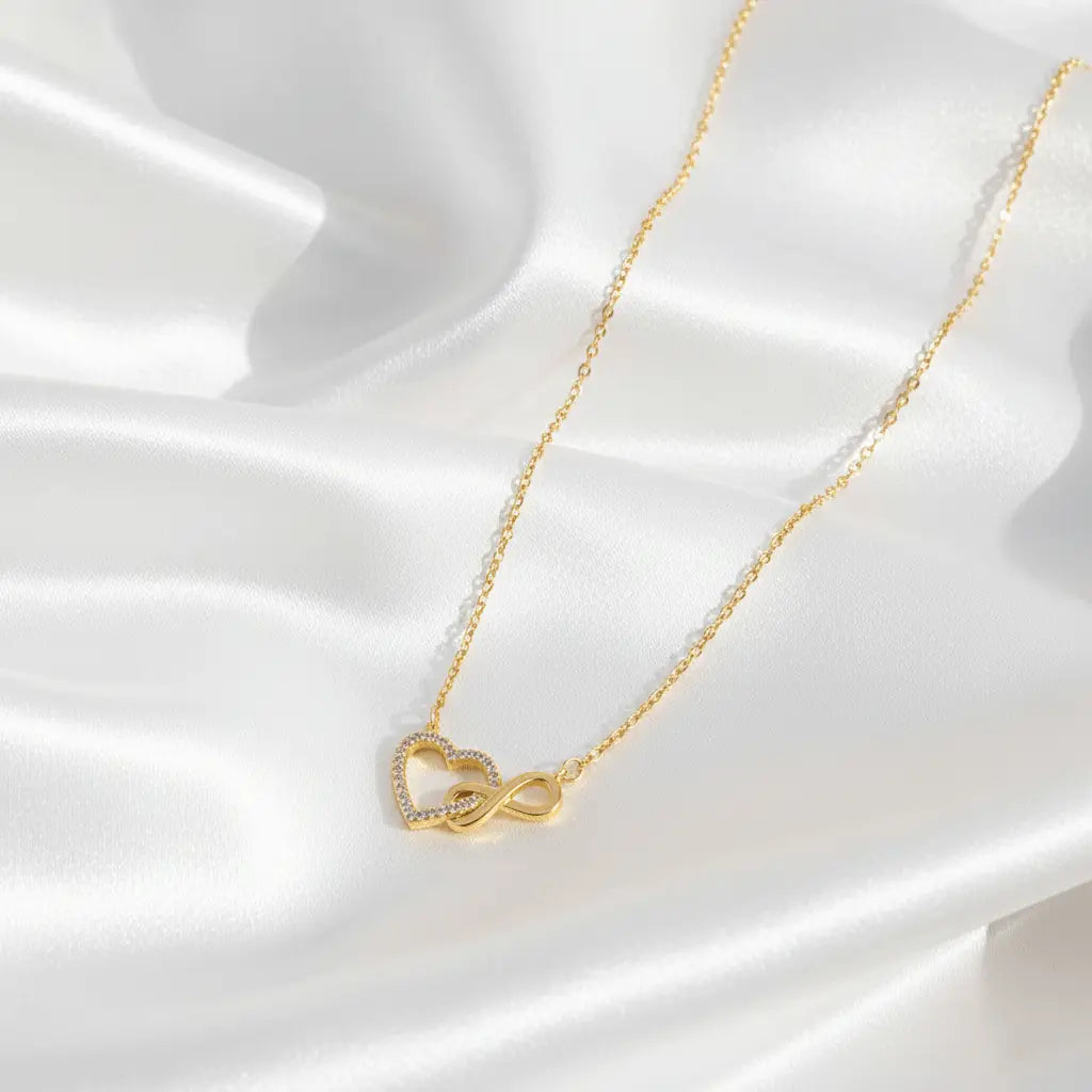 A delicate necklace with a heart-shaped pendant intertwined with an infinity symbol, accented with small crystals, displayed on smooth white satin fabric.
