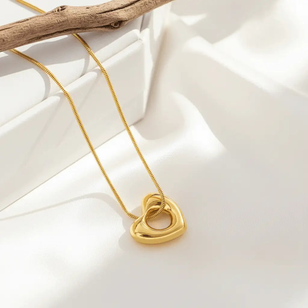 A polished gold heart-shaped pendant with a hollow center hanging from a delicate gold chain, displayed on a soft, elegant surface.