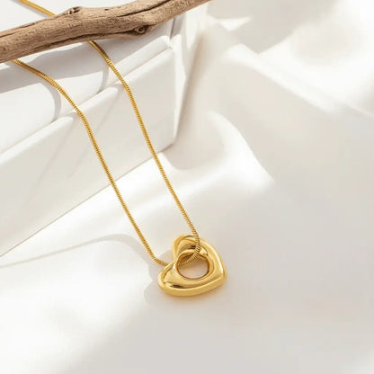 A polished gold heart-shaped pendant with a hollow center hanging from a delicate gold chain, displayed on a soft, elegant surface.