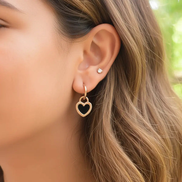 Woman wearing gold heart-shaped hoop earrings with black enamel center, close-up ear shot against a natural outdoor background.