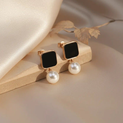 Gold-framed black square stud earrings with white pearl drops, styled on a neutral satin background for a luxury look.