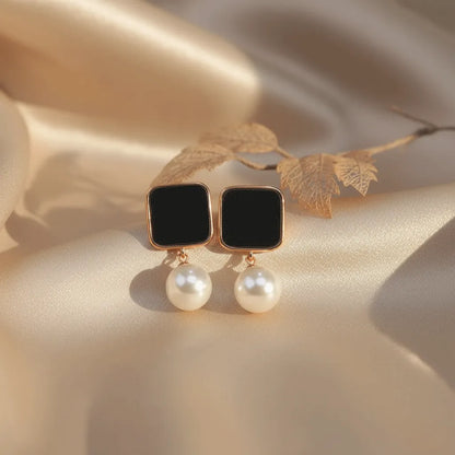 Elegant square black-stone stud earrings with a single dangling white pearl, displayed on soft champagne satin fabric.