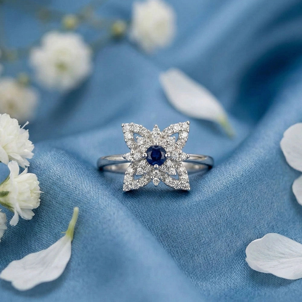 Silver AD ring with deep blue center stone and crystal detailing, styled on soft blue satin with white flower accents.