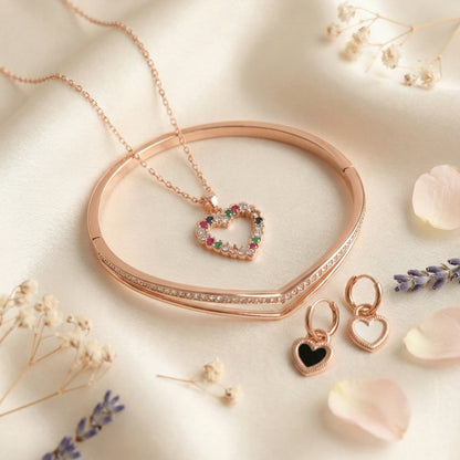 Rose gold jewelry set featuring a heart-shaped multicolor stone pendant necklace, matching heart earrings, and a sleek rose gold bangle arranged on a soft blush satin background with floral accents.