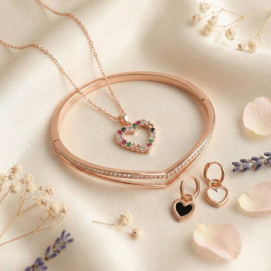 Rose gold jewelry set featuring a heart-shaped multicolor stone pendant necklace, matching heart earrings, and a sleek rose gold bangle arranged on a soft blush satin background with floral accents.