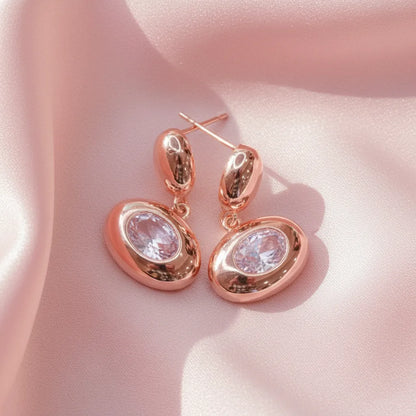 Close-up of Blush Glow Earrings with rose-gold finish and sparkling stones on a pink satin surface.