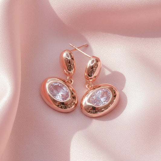 Close-up of Blush Glow Earrings with rose-gold finish and sparkling stones on a pink satin surface.