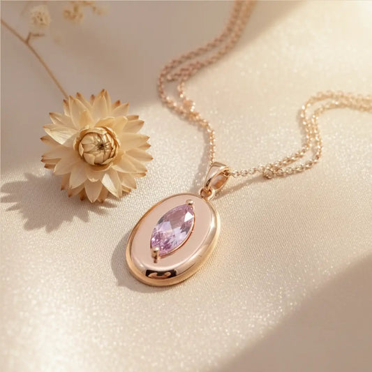 Rose gold necklace with a marquise-cut pink gemstone pendant placed on soft satin fabric beside a dried flower.