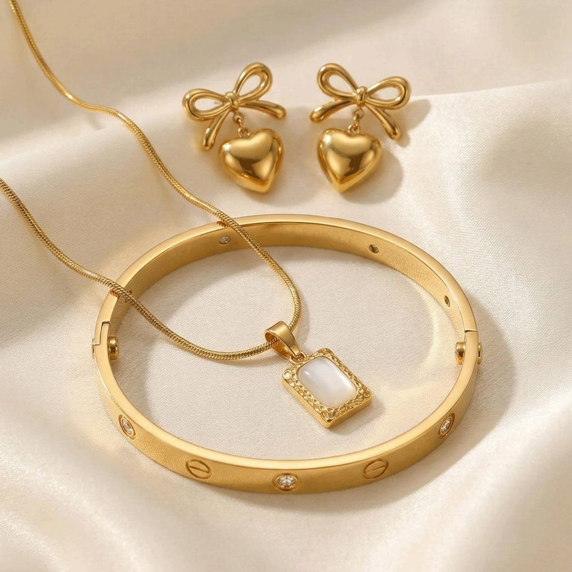 Gold jewelry set with a bangle, pendant necklace, and bow-shaped heart earrings on a soft light fabric background.