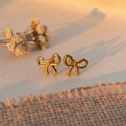 Gold bow-knot stud earrings placed on a soft beige fabric with warm sunlight and dried flowers.
