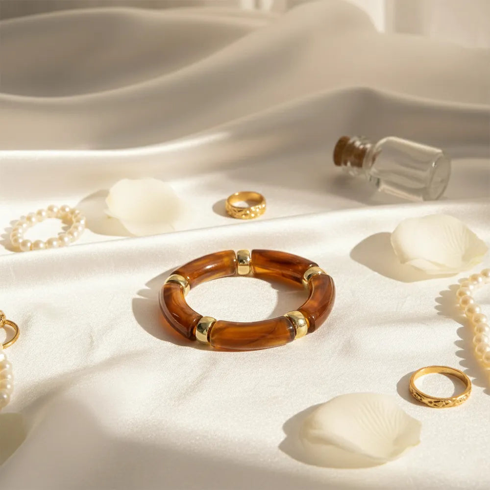 Brown resin bracelet with gold accents displayed on white satin with petals, pearls, and rings.