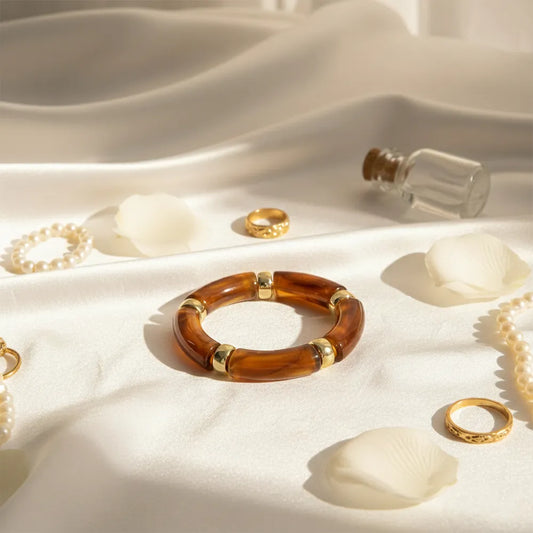 Brown resin bracelet with gold accents displayed on white satin with petals, pearls, and rings.