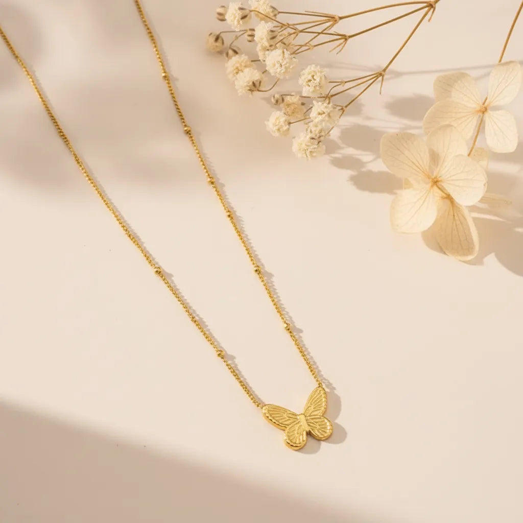 Delicate gold butterfly pendant necklace on a beaded chain with dried flowers in the background.