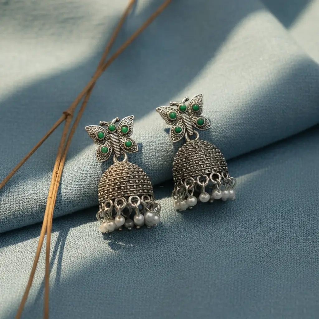 Silver butterfly jhumka earrings with green stones and pearl danglings displayed on a soft pastel blue fabric background.
