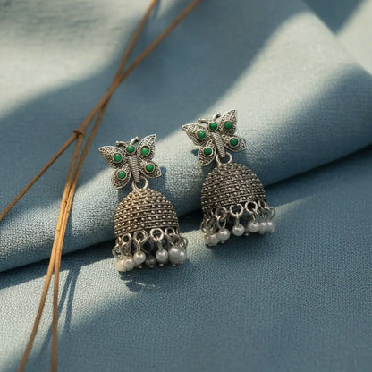 Silver butterfly jhumka earrings with green stones and pearl danglings displayed on a soft pastel blue fabric background.