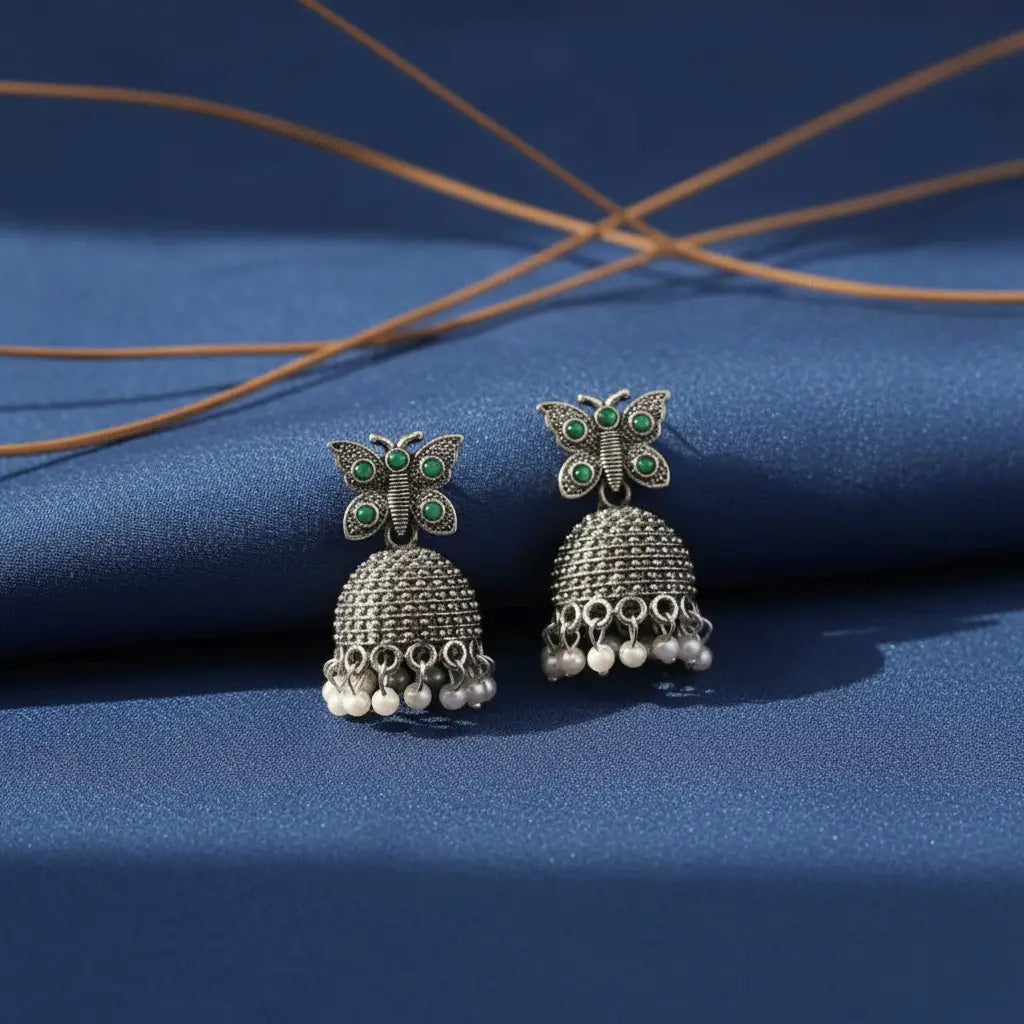Silver butterfly jhumka earrings with green stones and pearl drops displayed on a blue fabric background.”