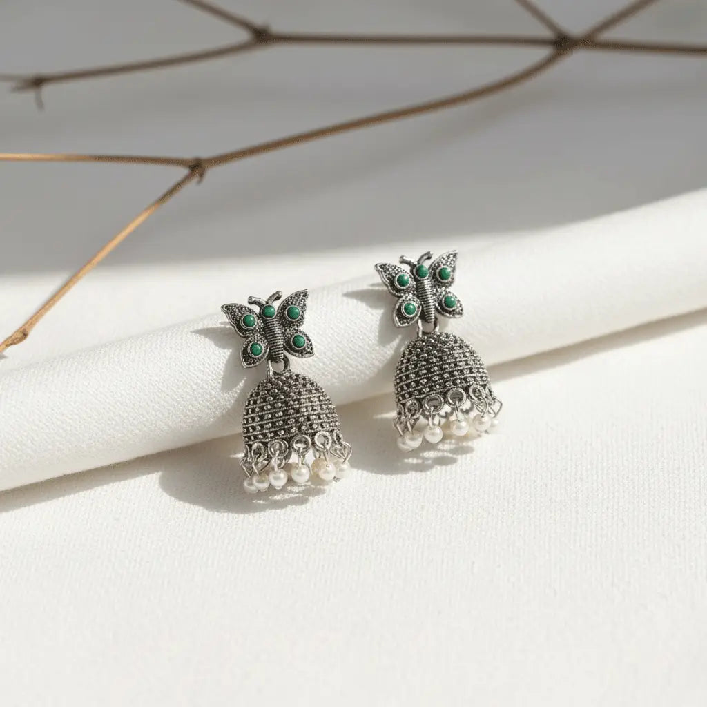 Silver butterfly jhumka earrings with green stones and pearl drops placed on a white textured background.