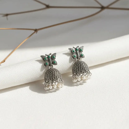 Silver butterfly jhumka earrings with green stones and pearl drops placed on a white textured background.