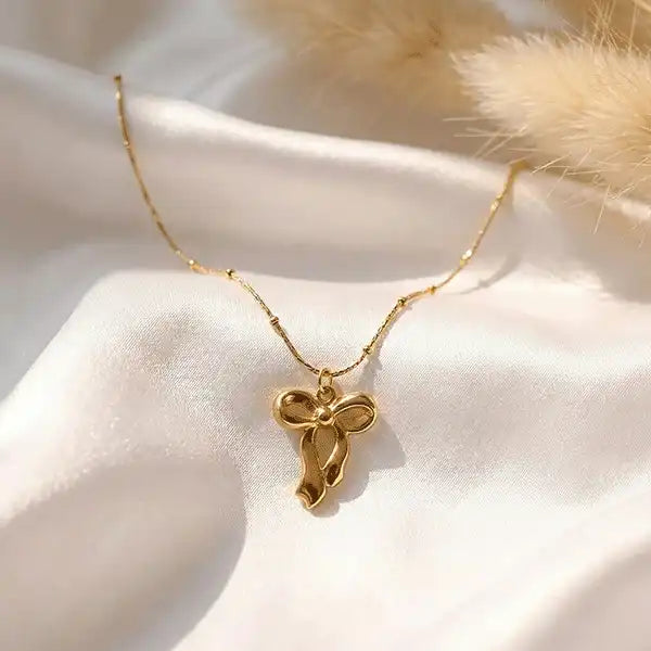 Classic bow drop pendant necklace in gold tone placed on white satin fabric with soft sunlight