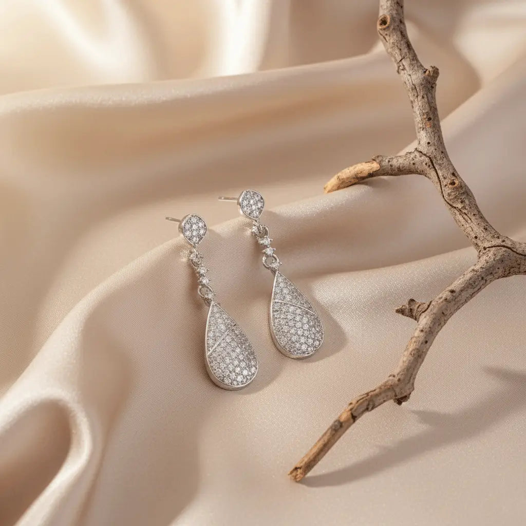 Elegant silver teardrop AD earrings with sparkling stones, displayed on champagne satin fabric beside a wooden twig.