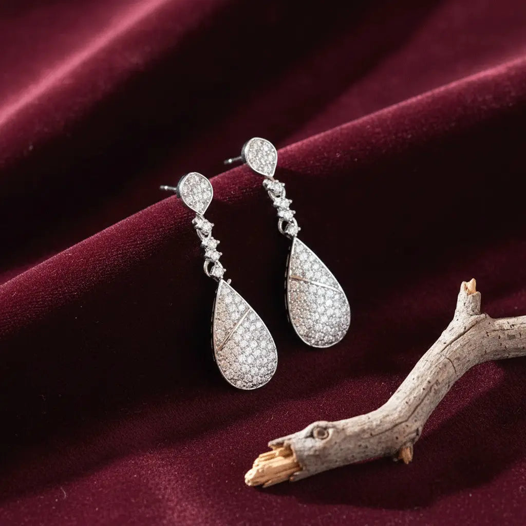 Sparkling silver teardrop AD earrings placed on rich maroon velvet fabric with a decorative wooden twig.