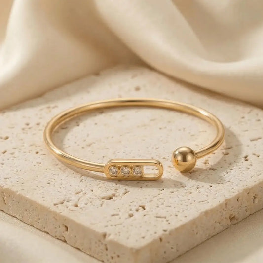 Minimalist gold open cuff bangle with three small crystals set in a rectangular bar, placed on a textured stone surface with soft neutral fabric in the background.