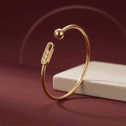 Gold open cuff bracelet with a polished ball end and a crystal-accented bar detail, displayed upright against a deep red background.