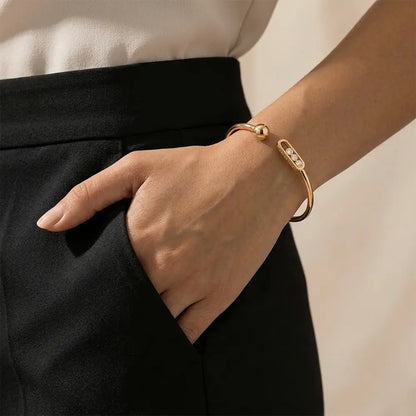 Woman wearing a sleek gold open cuff bangle with crystal accents on her wrist, paired with black pants and a white top, hand resting in pocket.