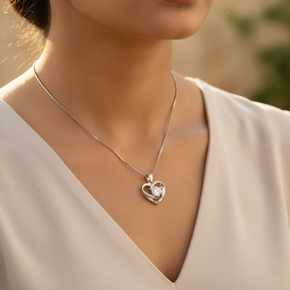 Crystal Heart AD Pendant necklace, a silver heart-shaped design with a brilliant central stone, styled on wood with flowers.