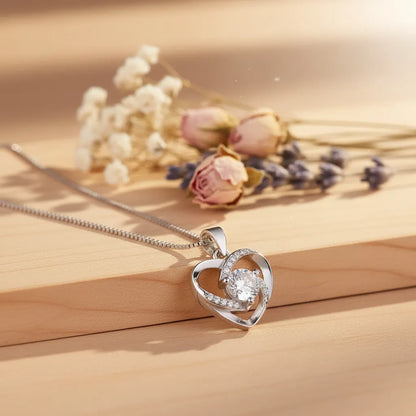 Crystal Heart AD Pendant necklace, a silver heart-shaped design with a brilliant central stone, styled on wood with flowers.