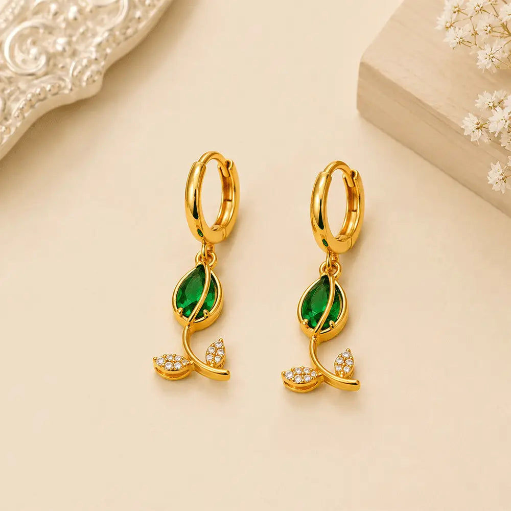 Pair of Emerald Petal anti tarnish hoop earrings with emerald green marquise-cut stones and crystal leaf motifs on a beige background.