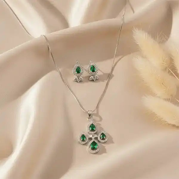 Emerald teardrop pendant and matching earrings set on beige satin fabric with soft sunlight and pampas grass