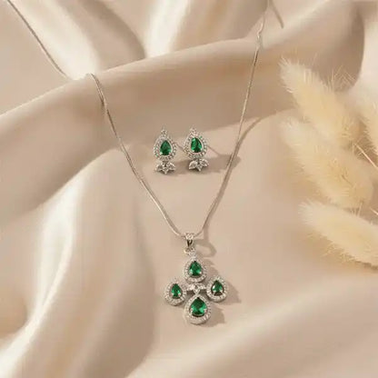 Emerald teardrop pendant and matching earrings set on beige satin fabric with soft sunlight and pampas grass