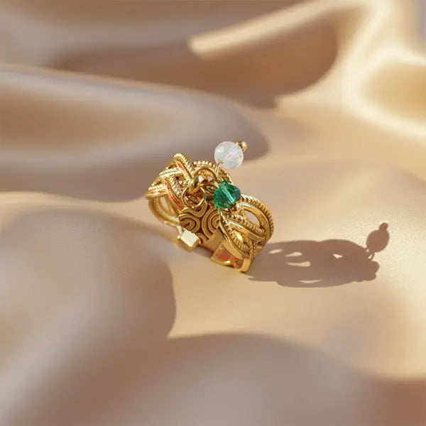 Ornate gold ring featuring detailed patterns and emerald-green charm resting on golden satin fabric with dramatic shadows.