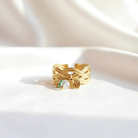 Gold adjustable ring with intricate engraved design and dangling clear and green crystal charms displayed on soft white satin.