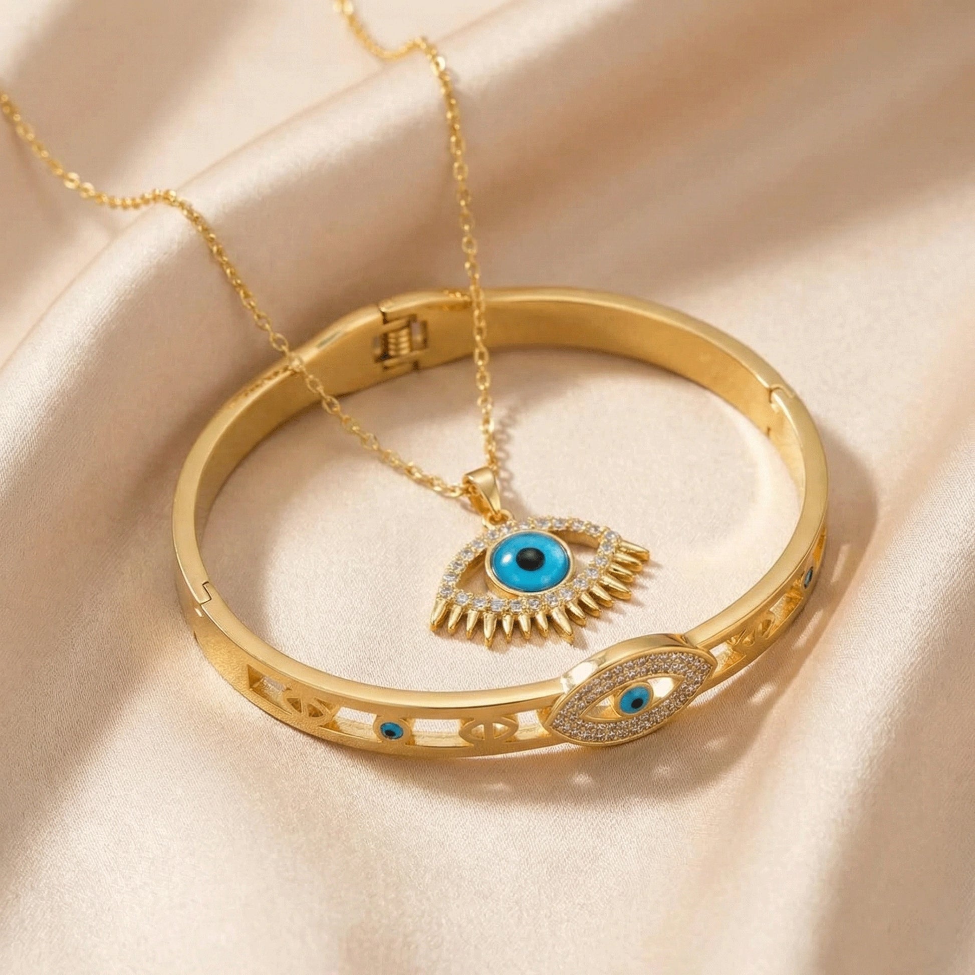 Gold evil eye jewelry set featuring a blue eye pendant necklace with crystal detailing and a matching gold bangle with eye motifs, styled on soft champagne satin fabric.