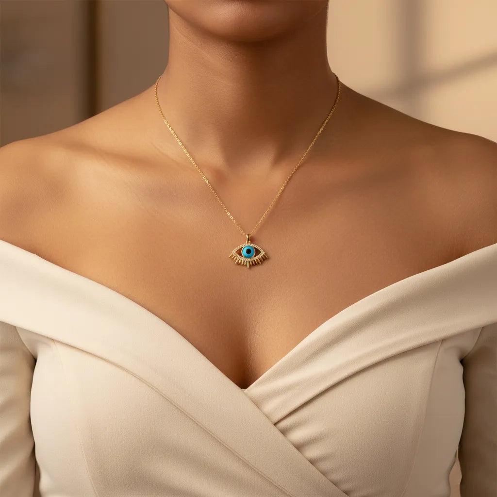Gold Evil Eye pendant necklace with a turquoise blue center and crystal-studded lashes, resting on dried wheat stalks.