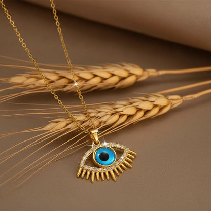 Gold Evil Eye pendant necklace with a turquoise blue center and crystal-studded lashes, resting on dried wheat stalks.