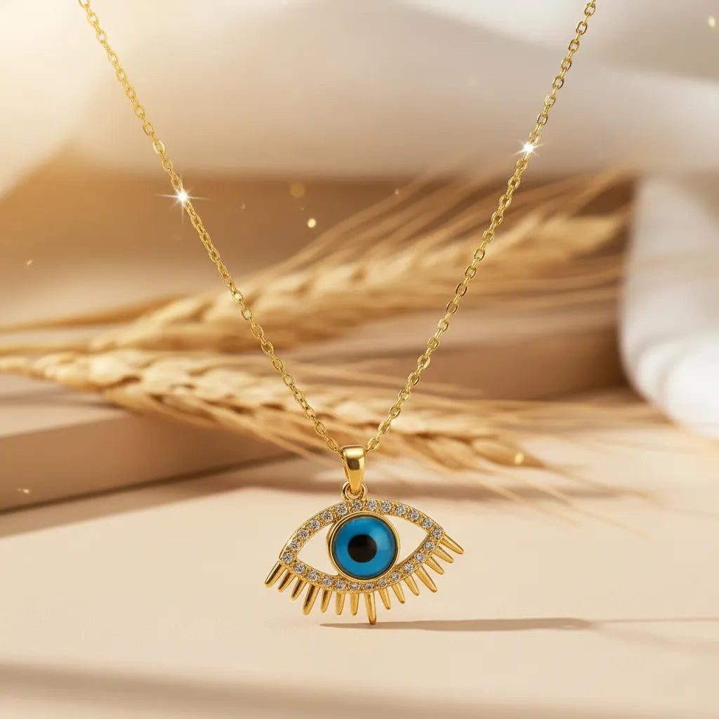Gold Evil Eye pendant necklace with a turquoise blue center and crystal-studded lashes, resting on dried wheat stalks.