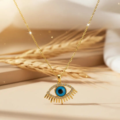 Gold Evil Eye pendant necklace with a turquoise blue center and crystal-studded lashes, resting on dried wheat stalks.