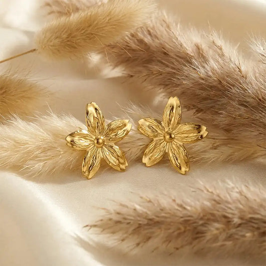 Gold flower statement stud earrings with textured petals displayed on white satin fabric with soft pampas grass background.