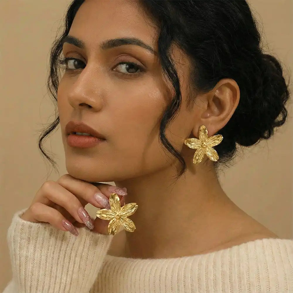 Model wearing gold flower statement stud earrings, showcasing bold floral design with a minimal makeup look and neutral background.