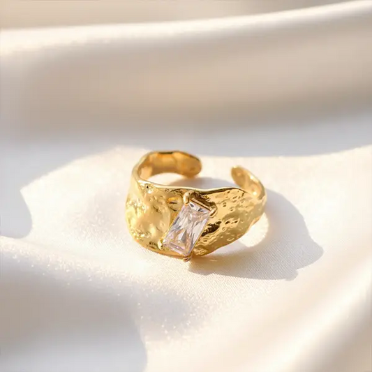 Gold textured open ring with a rectangular clear crystal displayed on soft satin fabric in warm sunlight.