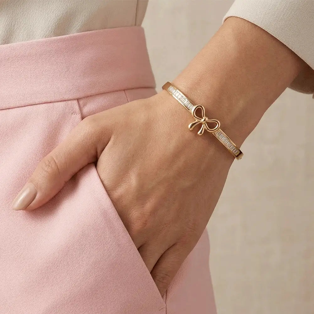 Woman wearing a slim gold bracelet with a bow centerpiece and baguette-cut crystal accents, paired with a white top and pink pants, hand in pocket.